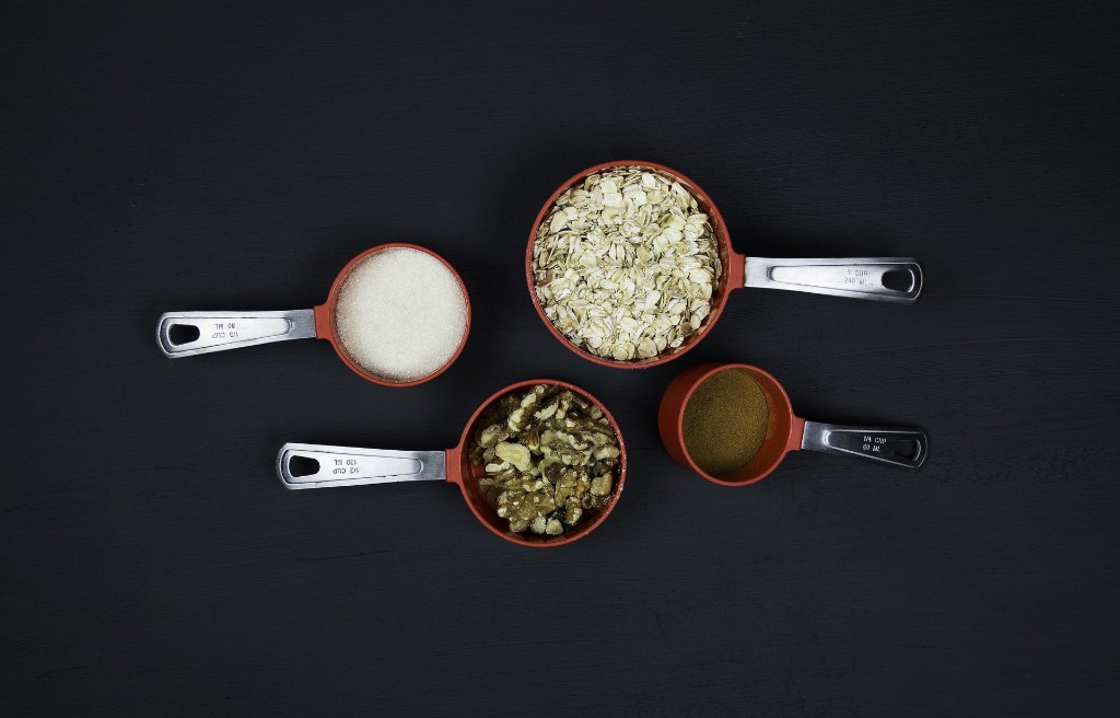 Measuring cups filled with oats, sugar, walnuts, and spice on a dark countertop